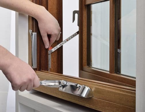 Hands measuring a wooden window frame with a tape measure and window crank.