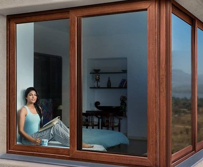 A woman sits comfortably on a wide wooden window sill, reading a book and holding a mug.