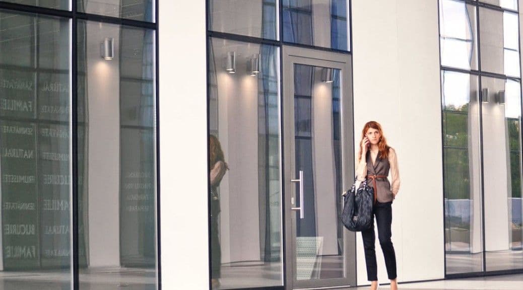 A woman stands outside a modern glass building, holding a black bag.