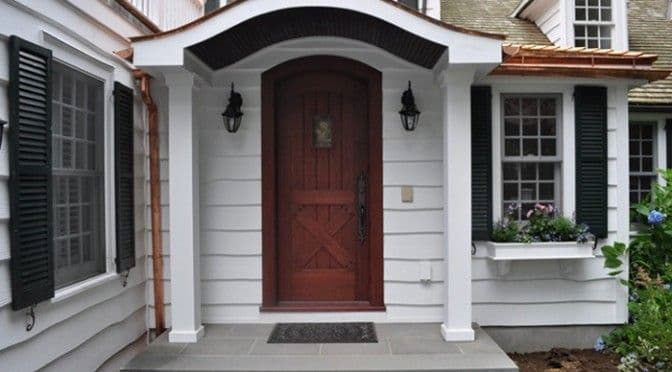 Brown wooden front door with arched frame on a white clapboard house.