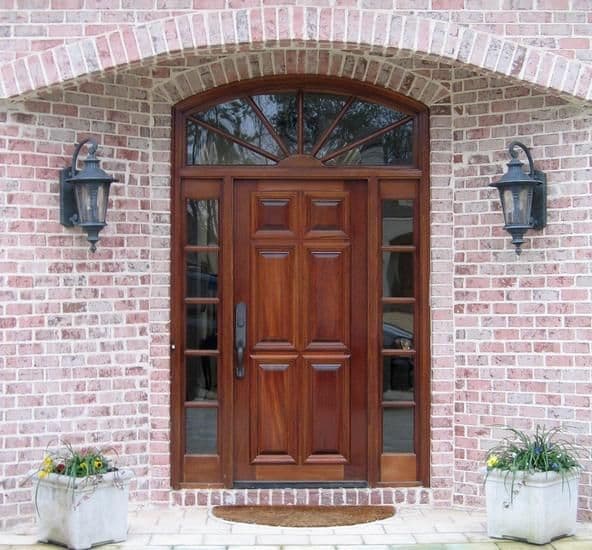 Elegant dark wooden front door framed by a light red brick archway with two ornate sconces.