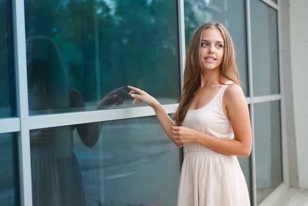 A young woman in a light dress stands smiling by a modern glass building.