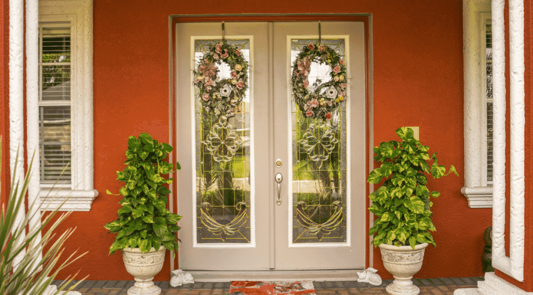 Reddish-orange house entrance with double glass doors, wreaths, and potted plants.