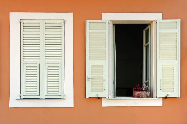 Two windows with white louvered shutters on an orange wall, one is open.