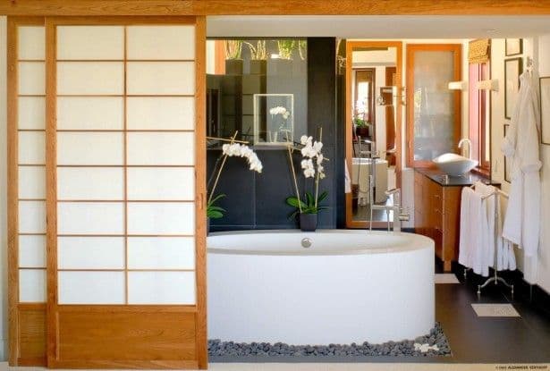 A modern bathroom featuring a round white tub, shoji screen, dark tiles, and orchid plants.