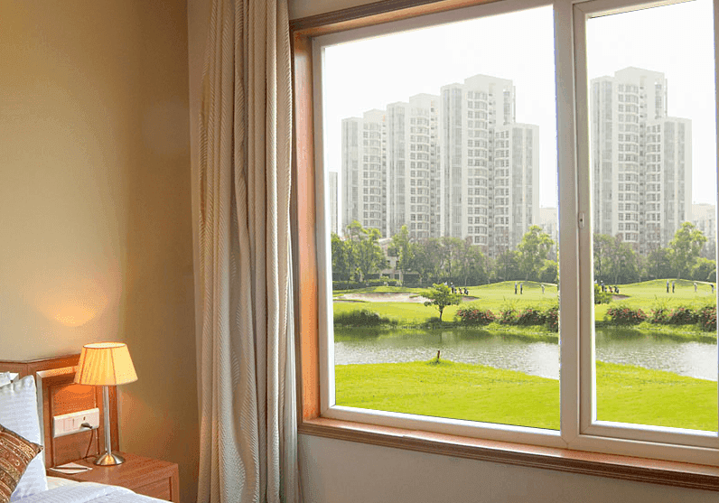 Room window view of modern buildings, a green park, and a serene lake.