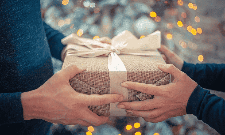 Hands exchanging a holiday gift wrapped in brown paper with a ribbon and festive lights.