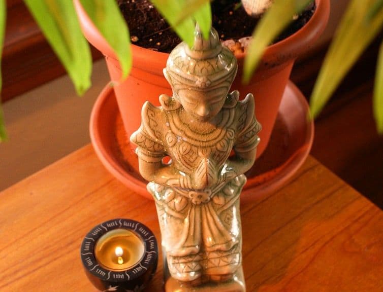 Light-colored statue in prayer pose, small lit candle, and potted plant on a wooden surface.