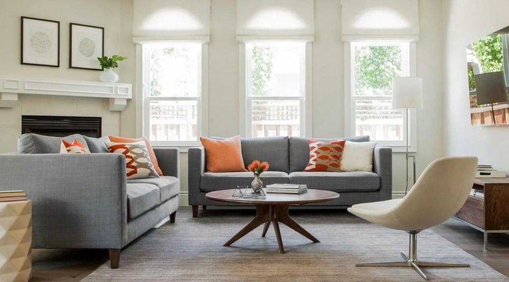 Modern living room with gray sofas, orange pillows, a wooden coffee table, and windows.