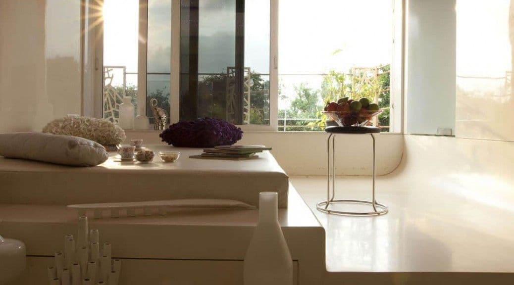 Sunny minimalist living room with white platform furniture, decorative pillows, and large windows.