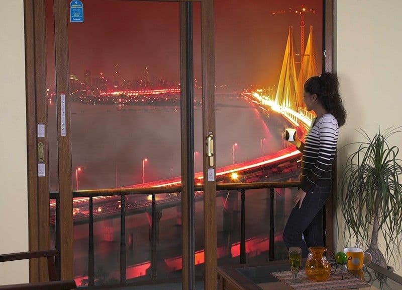 A woman stands on a balcony looking at a brightly lit city bridge at night.