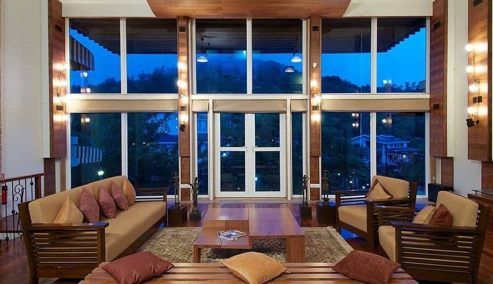 A modern living room with large windows, wooden furniture, light cushions, and ambient wall lighting.